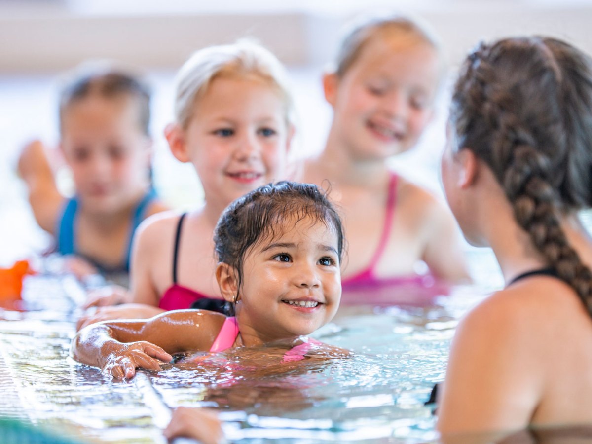 Gruppe von Schwimmschülerinnen im Becken. Die Schwimmtrainerin dreht der Kamera den Rücken zu.
