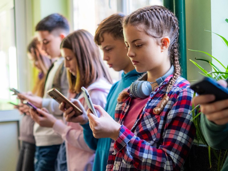 Gruppe Schüler*innen steht in einer Reihe am Fenster und jede*r schaut aufs eigene Handy.