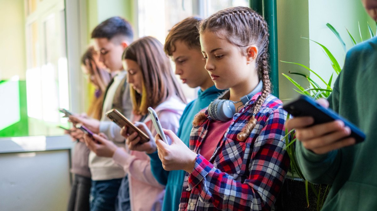 Gruppe Schüler*innen steht in einer Reihe am Fenster und jede*r schaut aufs eigene Handy.