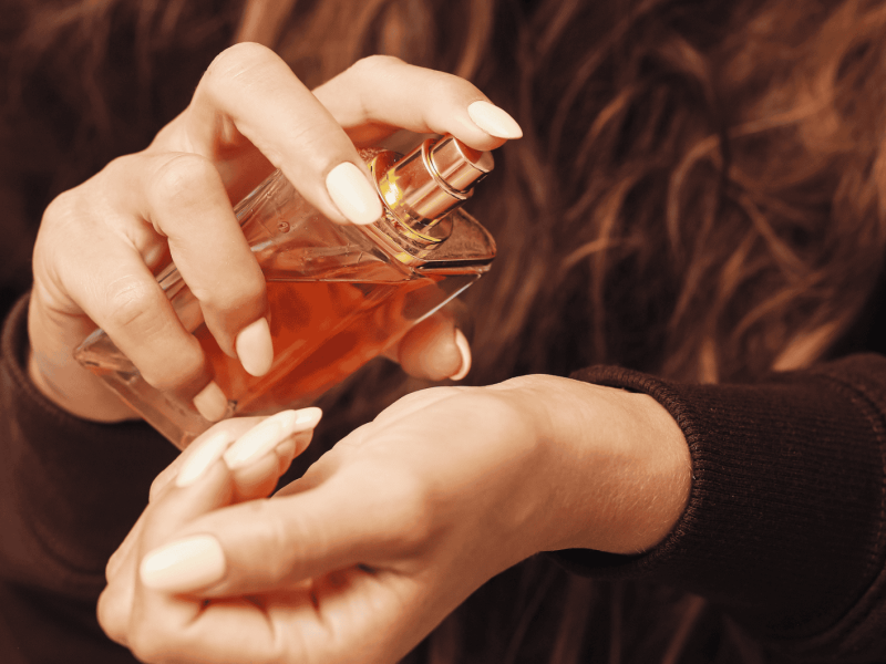 Frau in braunem Pullover sprüht Parfum auf Handgelenk.
