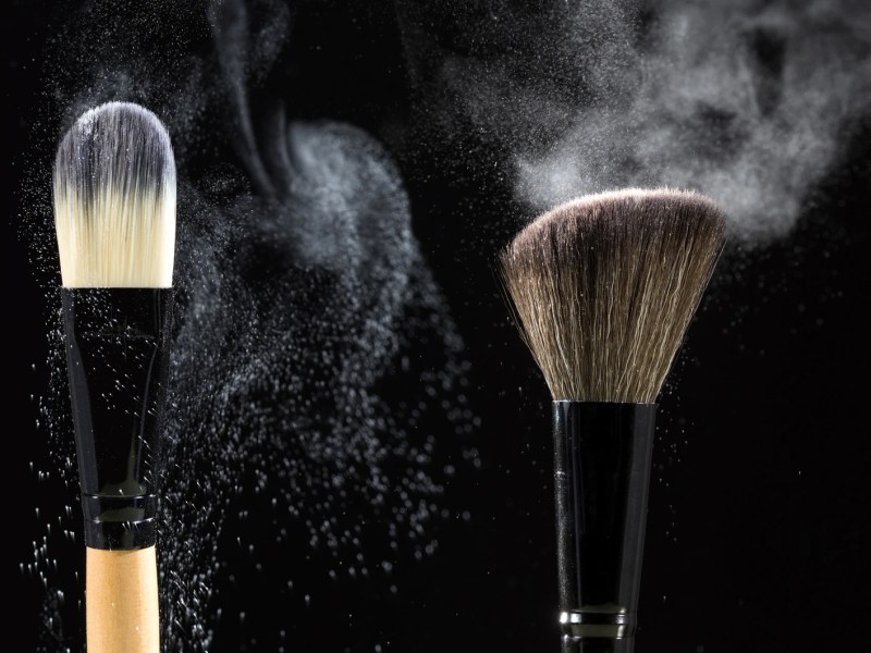 Zwei Make-up-Pinsel wirbeln feinen Puder in der Luft auf, vor schwarzem Hintergrund dramatisch beleuchtet.