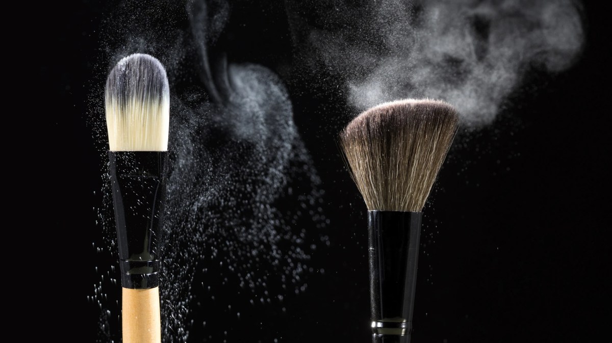 Zwei Make-up-Pinsel wirbeln feinen Puder in der Luft auf, vor schwarzem Hintergrund dramatisch beleuchtet.