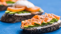 Sandwiches mit geräuchertem Lachs, Eiern, Kernen, Frischkäse und Avocado auf blauem Hintergrund.