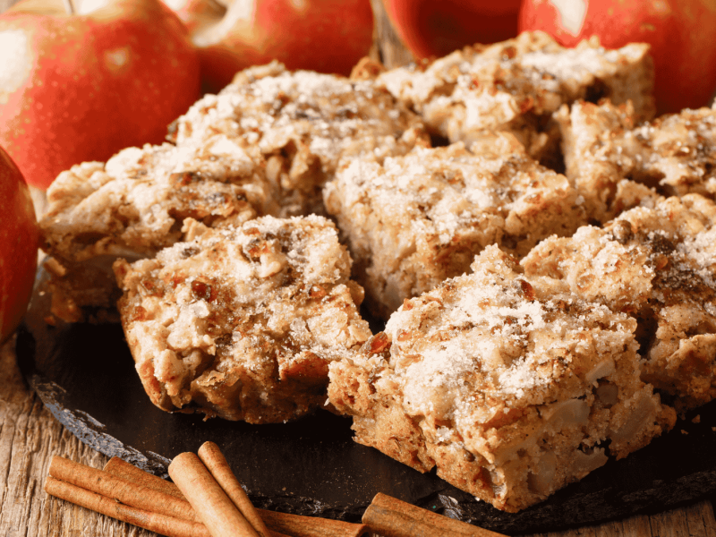 Apfel-Zimt-Kuchen auf einem Holztisch neben Äpfeln und Zimtstangen.