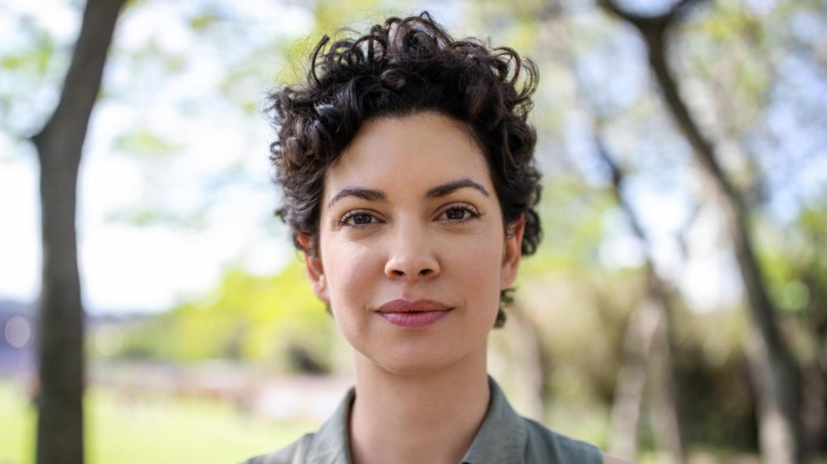 Junge Frau mit dunklen kurzer trendiger Locken-Frisur blickt selbstbewusst in die Kamera.