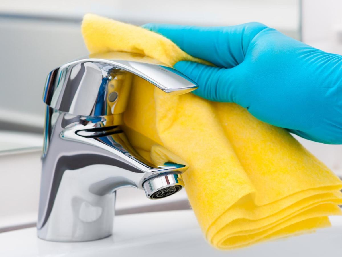 Eine Hand mit einem blauen Handschuh reinigt den Wasserhahn am Waschbecken mit einem gelben Tuch.