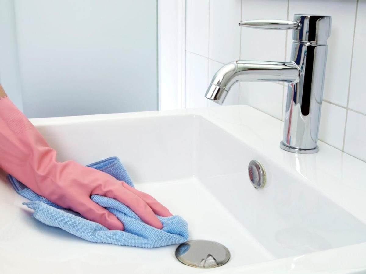 Frau mit einem rosa Handschuh macht das Waschbecken mit einem blauen Tuch sauber.