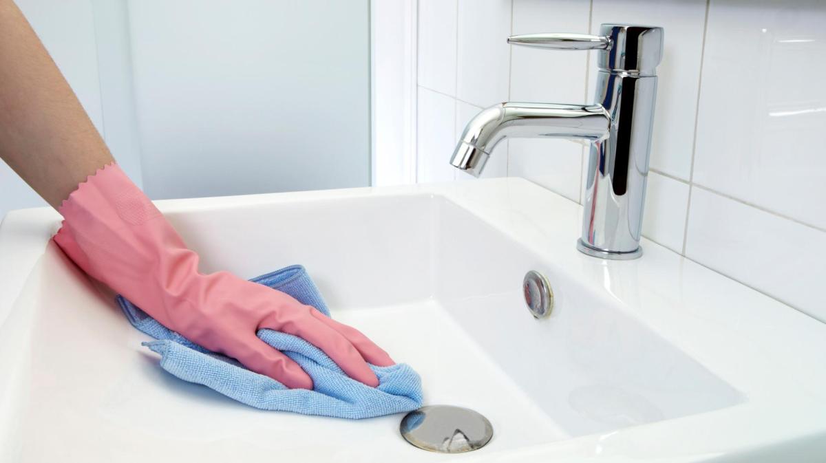 Frau mit einem rosa Handschuh macht das Waschbecken mit einem blauen Tuch sauber.