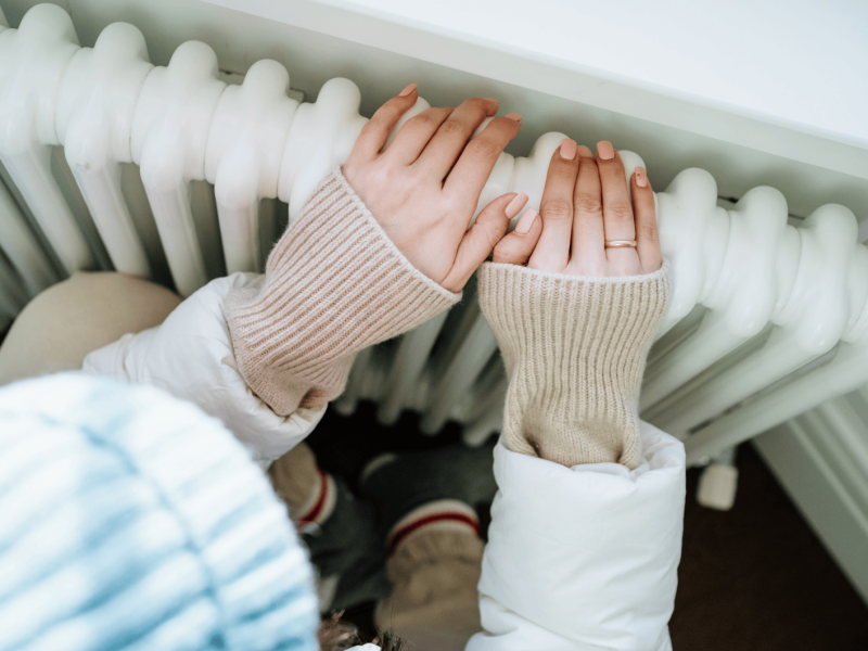 Frau sitzt warm angezogen vor der Heizung und hält ihre Hände auf die Heizung.