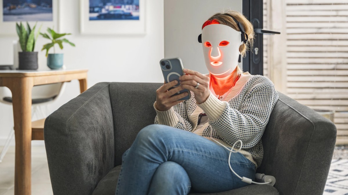 Frau nutzt LED-Gesichtsmaske zu Hause auf ihrem Sofa.