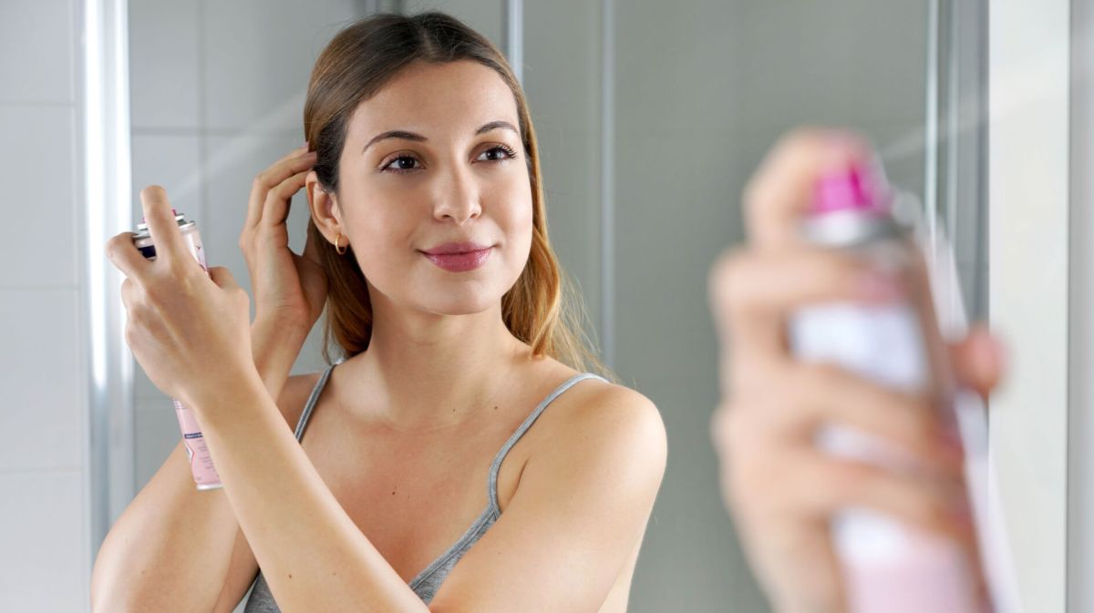 Junge Frau steht vor dem Spiegel und trägt Trockenshampoo auf ihre Haare auf.