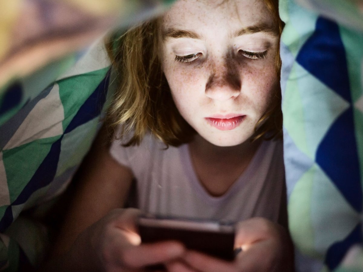 Mädchen im Teenageralter liegt im Bett unter der Decke und schaut auf ihr Handy.