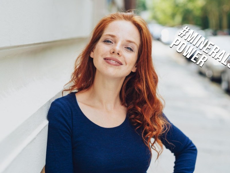 Hübsche Frau mit rotem Haar steht auf einem Gehweg und lächelt in die Kamera.