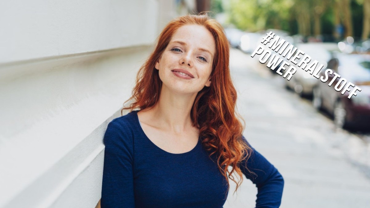 Hübsche Frau mit rotem Haar steht auf einem Gehweg und lächelt in die Kamera.