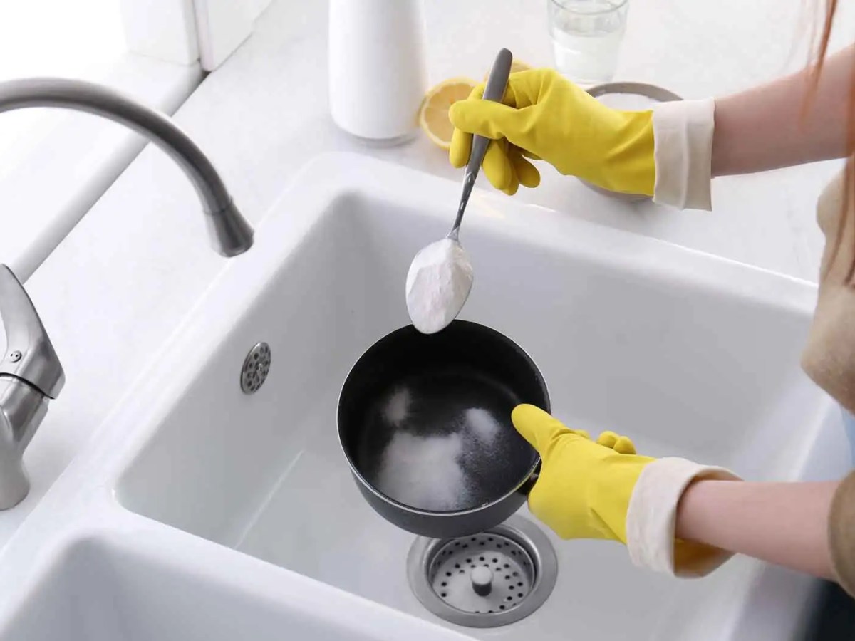 Frau mit Putzhandschuhen gibt Backpulver mit einem Essl&ouml;ffel in eine Sch&uuml;ssel &uuml;ber dem Waschbecken.