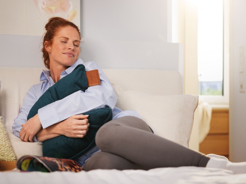 Frau sitzt mit einem Beurer Heizkissen auf dem Sofa.