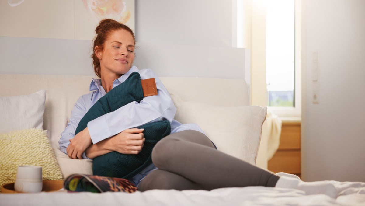 Frau sitzt mit einem Beurer Heizkissen auf dem Sofa.