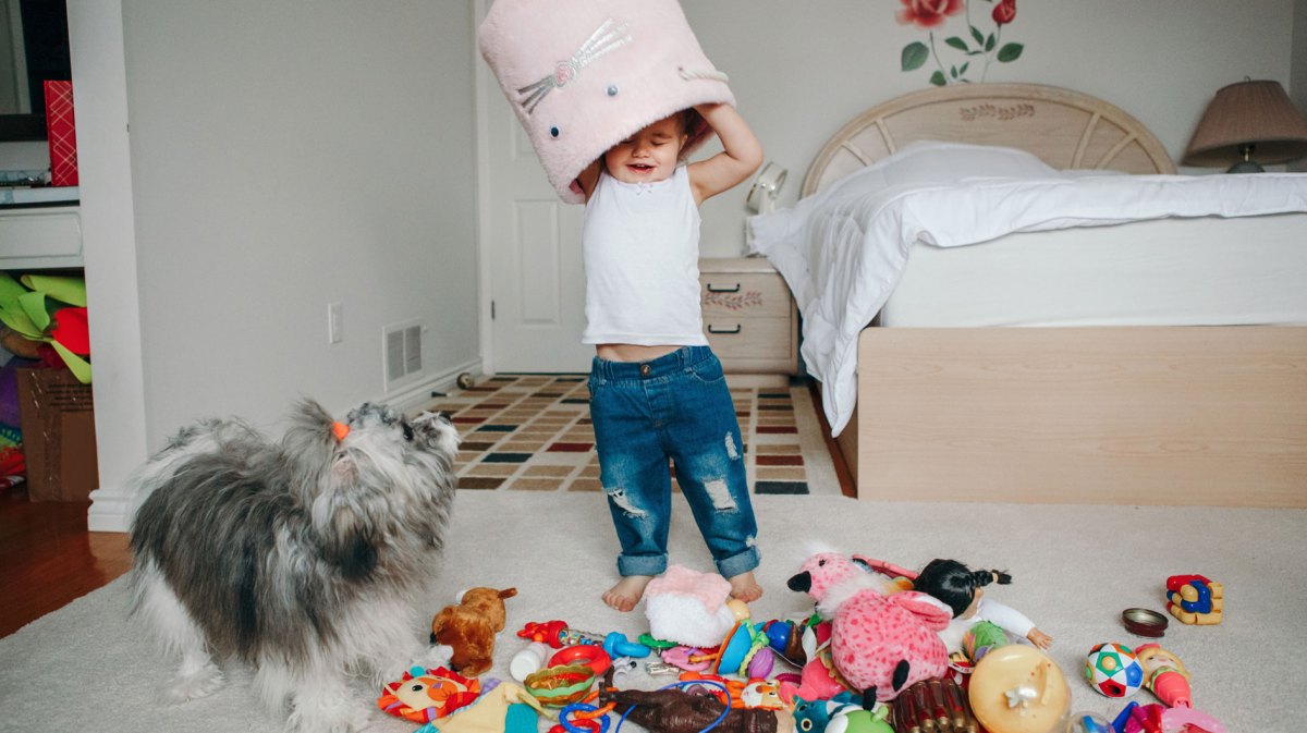 Kleinkind spielt im Zimmer und kippt einen Spielzeugkorb über seinem Kopf aus, sodass alles auf dem Boden liegt.