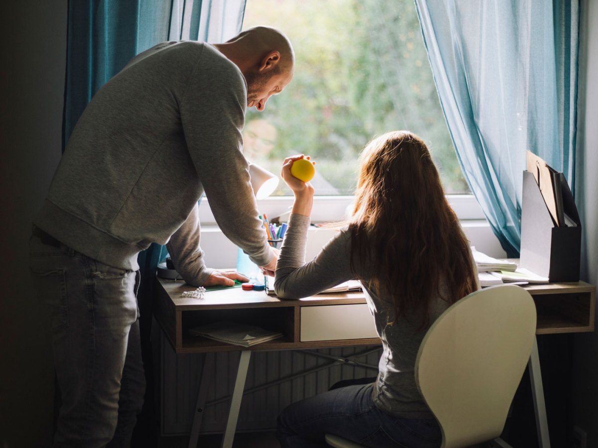 Teenager-Mädchen sitzt mit Stressball in der Hand am Schreibtisch, sein Vater steht daneben und erklärt etwas.