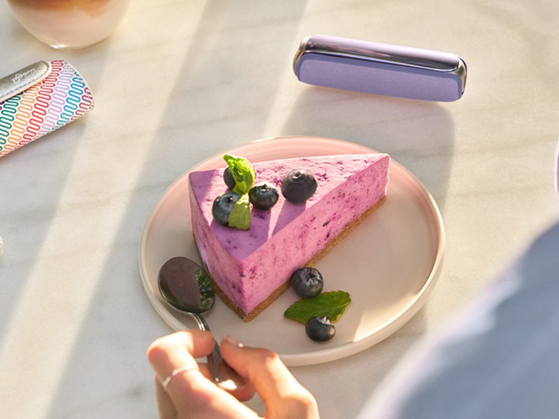 Hand mit silbernem Ring greift zu einem Löffel neben Blaubeerkuchen, bunte Serviette und violett-glänzendes Accessoire im Hintergrund.