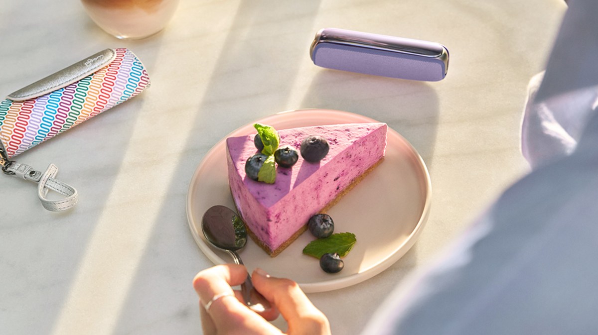 Hand mit silbernem Ring greift zu einem Löffel neben Blaubeerkuchen, bunte Serviette und violett-glänzendes Accessoire im Hintergrund.