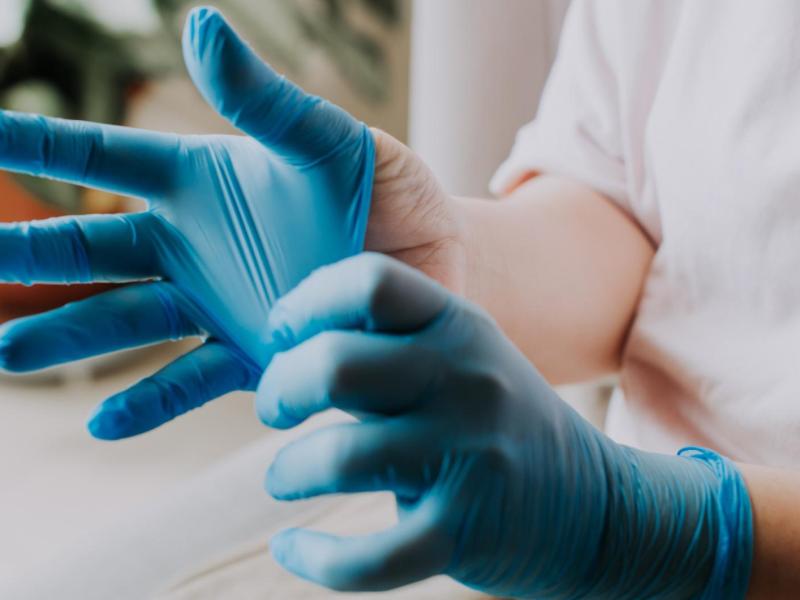 Eine Person mit hellem Shirt streift sich konzentriert blaue Einweghandschuhe über, im Hintergrund unscharf eine grüne Zimmerpflanze.