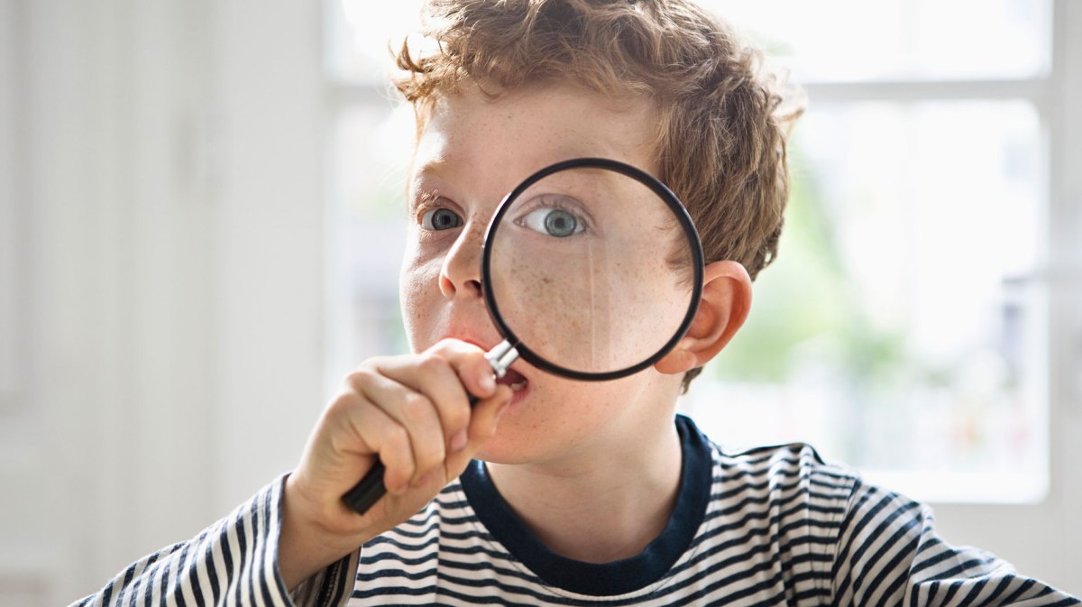 Porträt eines Jungen, ca. 8 Jahre, der sich eine Lupe vor sein linkes Auge hält.