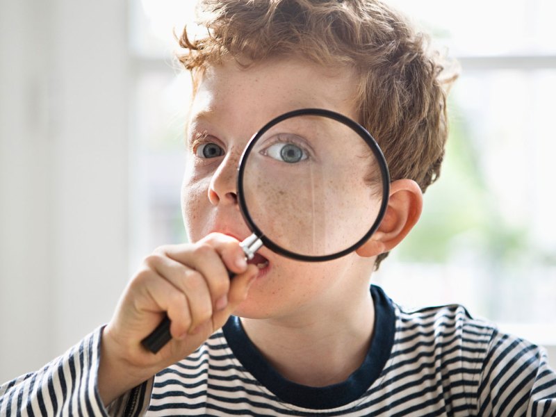 Porträt eines Jungen, ca. 8 Jahre, der sich eine Lupe vor sein linkes Auge hält.