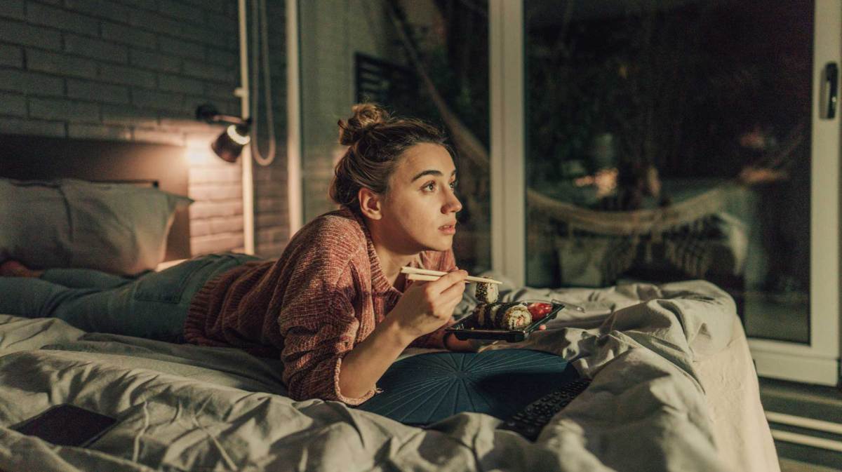 Frau liegt auf dem Bett, isst Sushi und schaut etwas Spannendes.