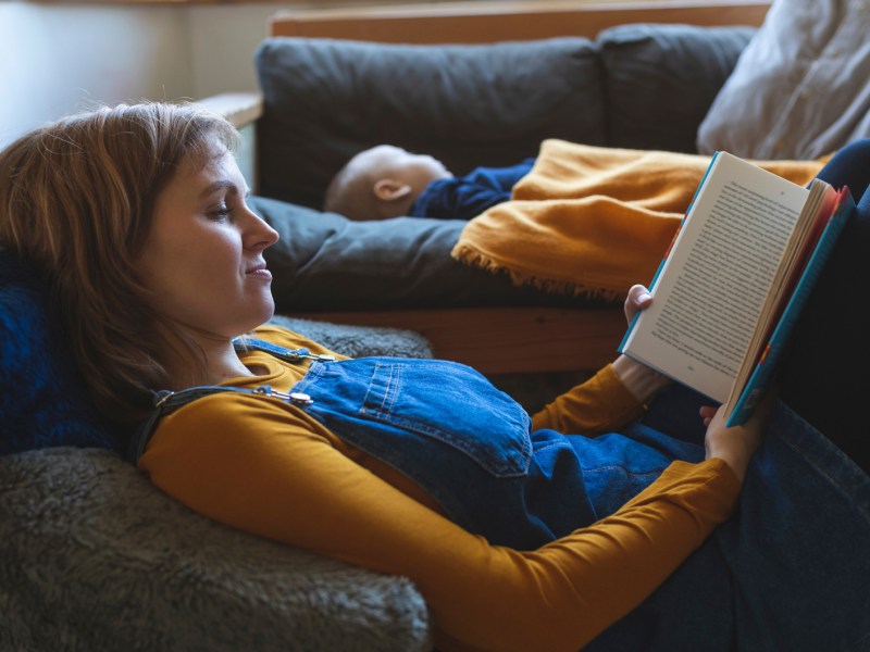 Mama liest Buch, während Kind im Hintergrund auf der Couch schläft.