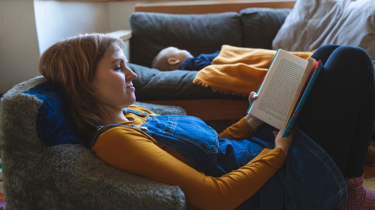 Mama liest Buch, während Kind im Hintergrund auf der Couch schläft.