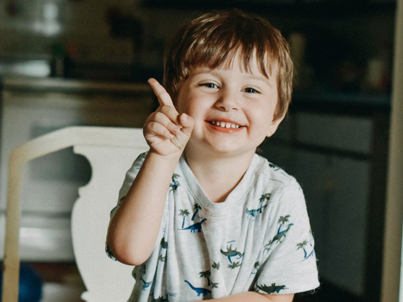 Kind am Küchentisch hebt den Finger und möchte etwas sagen.