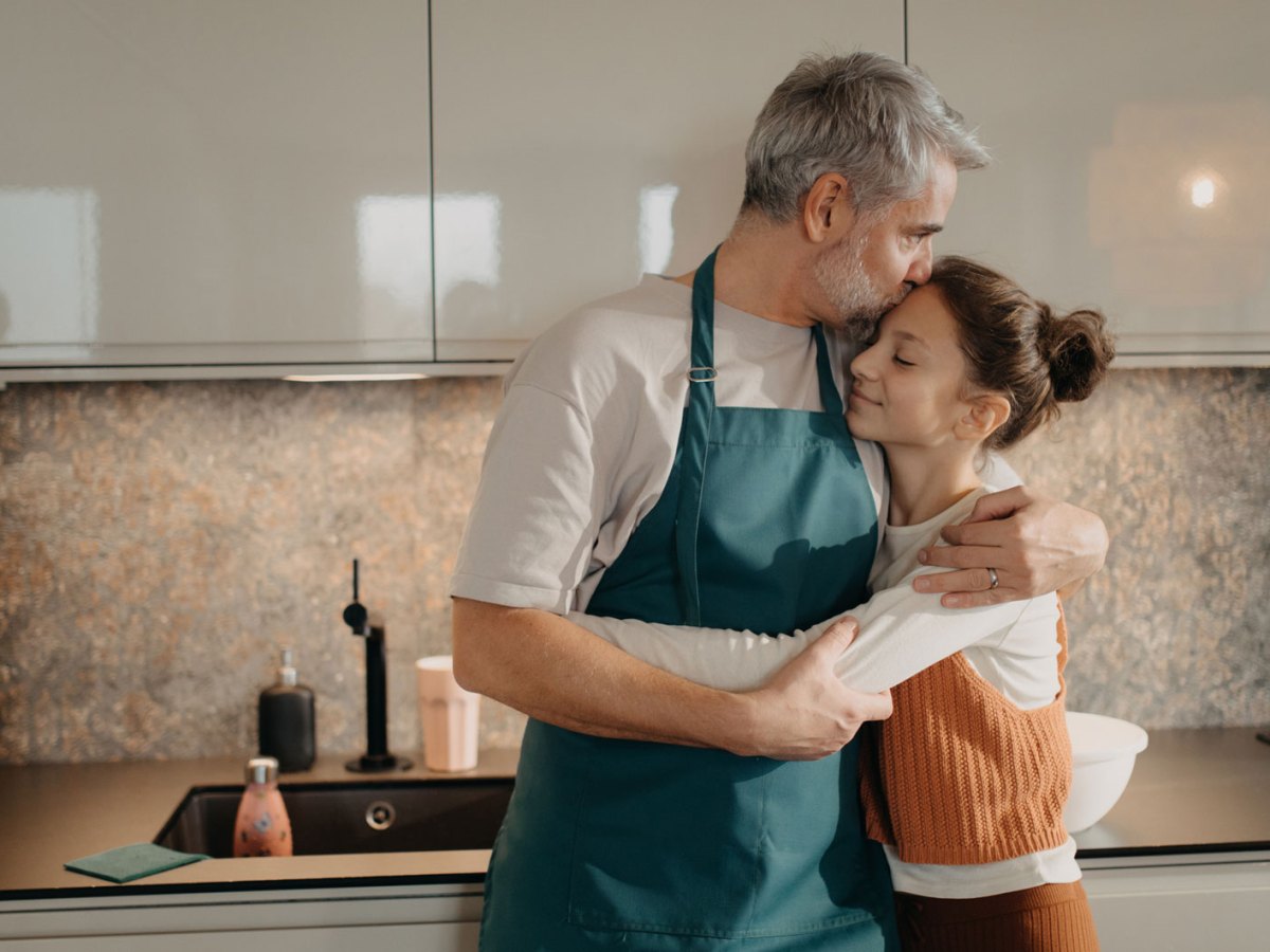 Vater tr&auml;gt eine Sch&uuml;rze, umarmt Tochter in der K&uuml;che und gibt ihr einen Kuss auf die Stirn.
