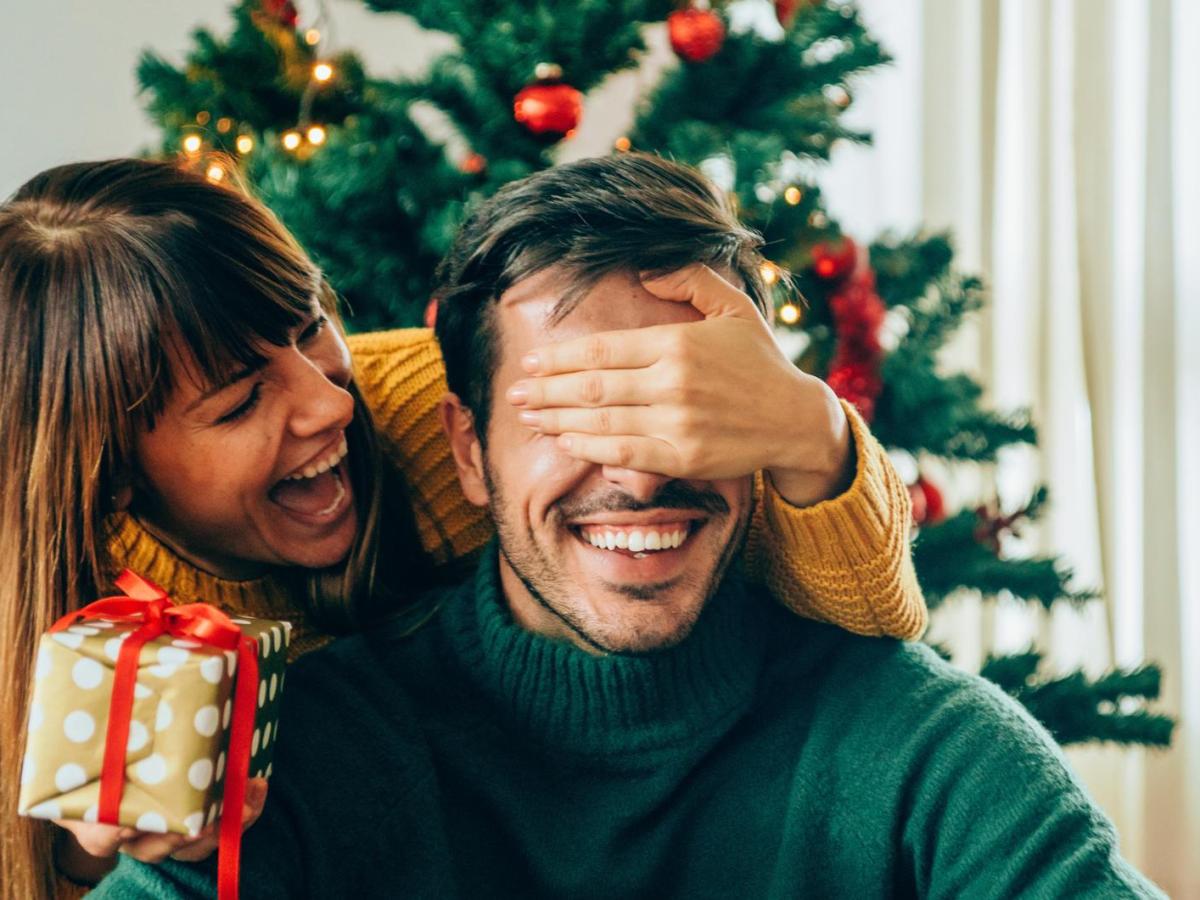 Frau h&auml;lt Mann vor dem Weihnachtsbaum die Hand vor die Augen, um ihn mit einem Geschenk zu &uuml;berraschen.
