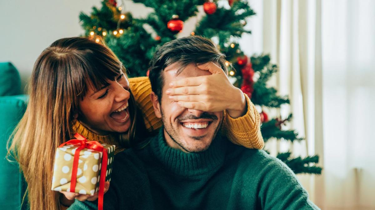 Frau hält Mann vor dem Weihnachtsbaum die Hand vor die Augen, um ihn mit einem Geschenk zu überraschen.