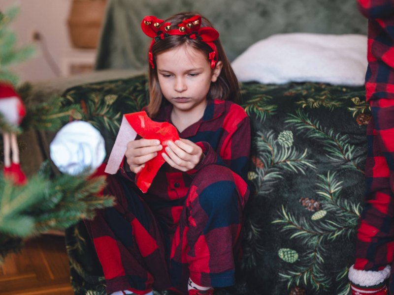 Mädchen sitzt am Weihnachtsmorgen enttäuscht neben dem Weihnachtsbaum und packt ein Geschenk aus.