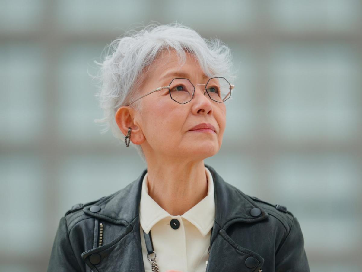 Junggebliebene ältere Frau mit weißen Haaren und Lederjacke und Brille und guckt zur Seite.