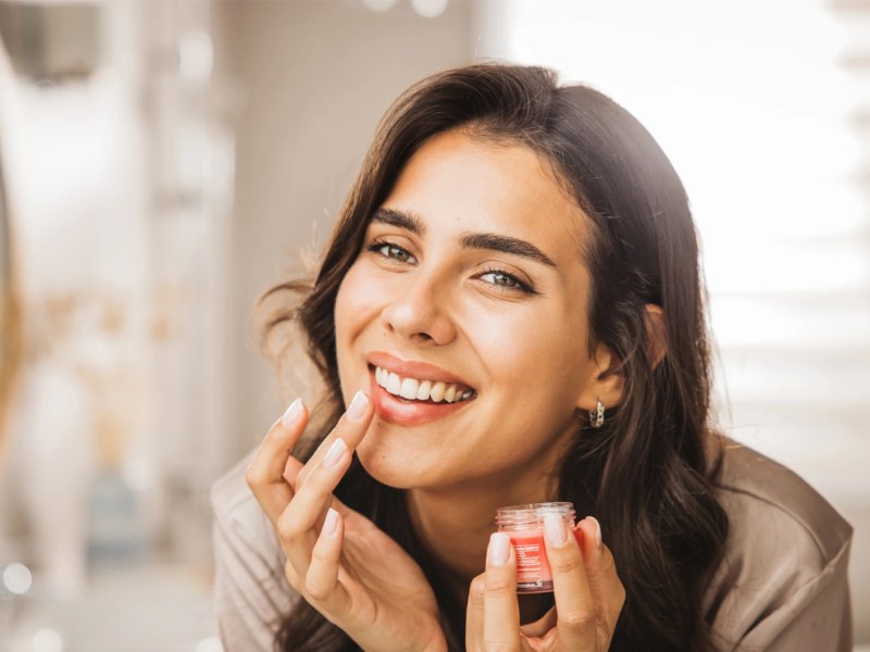 Frau vorm Spiegel, die sich Lippenpflege auf die Lippen aufträgt.
