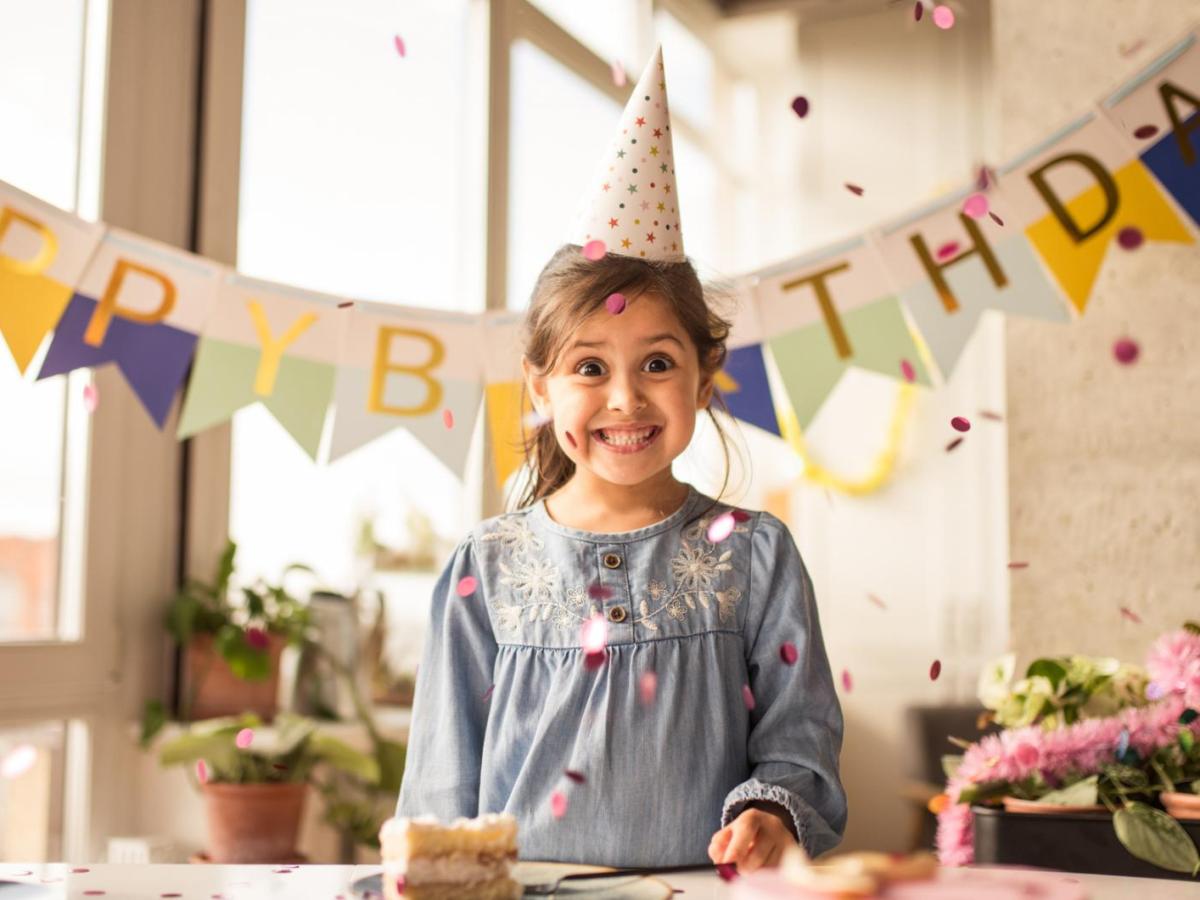 Mädchen hat Geburtstag und steht in einem geschmückten Wohnzimmer mit einem Partyhütchen auf dem Kopf und freut sich.
