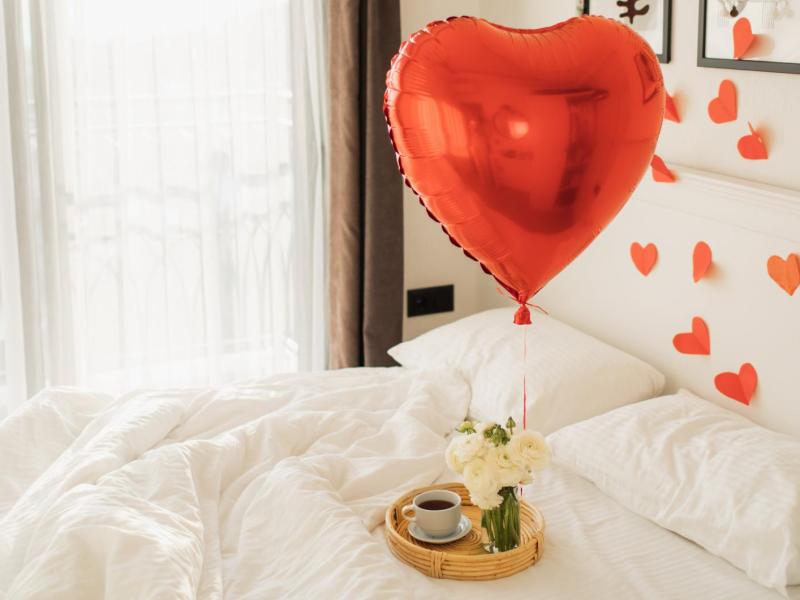 Roter Herzballon auf einem Bett mit weißer Bettwäsche, auf dem ein kleines Tablett mit einem Kaffee steht.