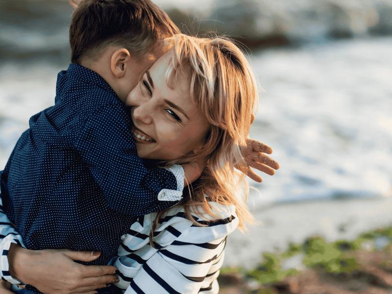 Frau mit blonden Haaren umarmt l&auml;chelnd einen kleinen Jungen.
