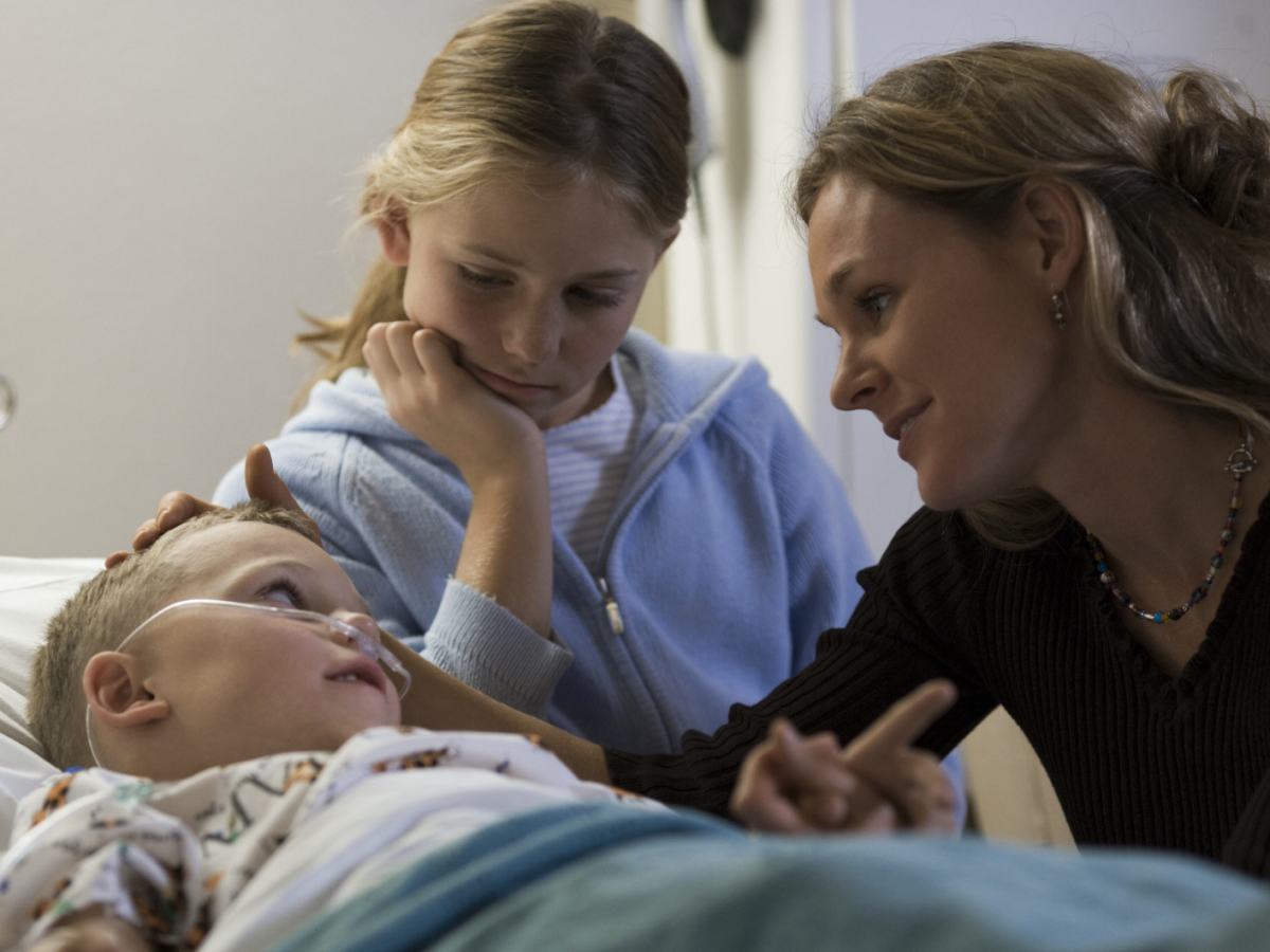 Mutter und älteres Geschwisterkind sitzen am Krankenbett eines Kleinkindes