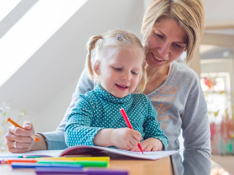 Junge Mutter sitzt mit ihrer Tochter auf dem Schoß am Tisch und malt.