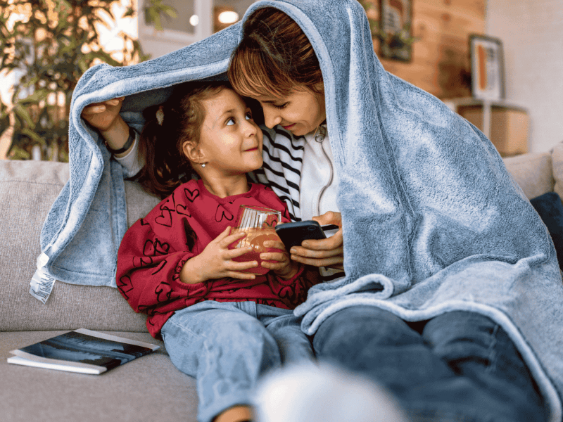 Mutter und Tochter kuscheln auf Sofa unter Kuscheldecke.