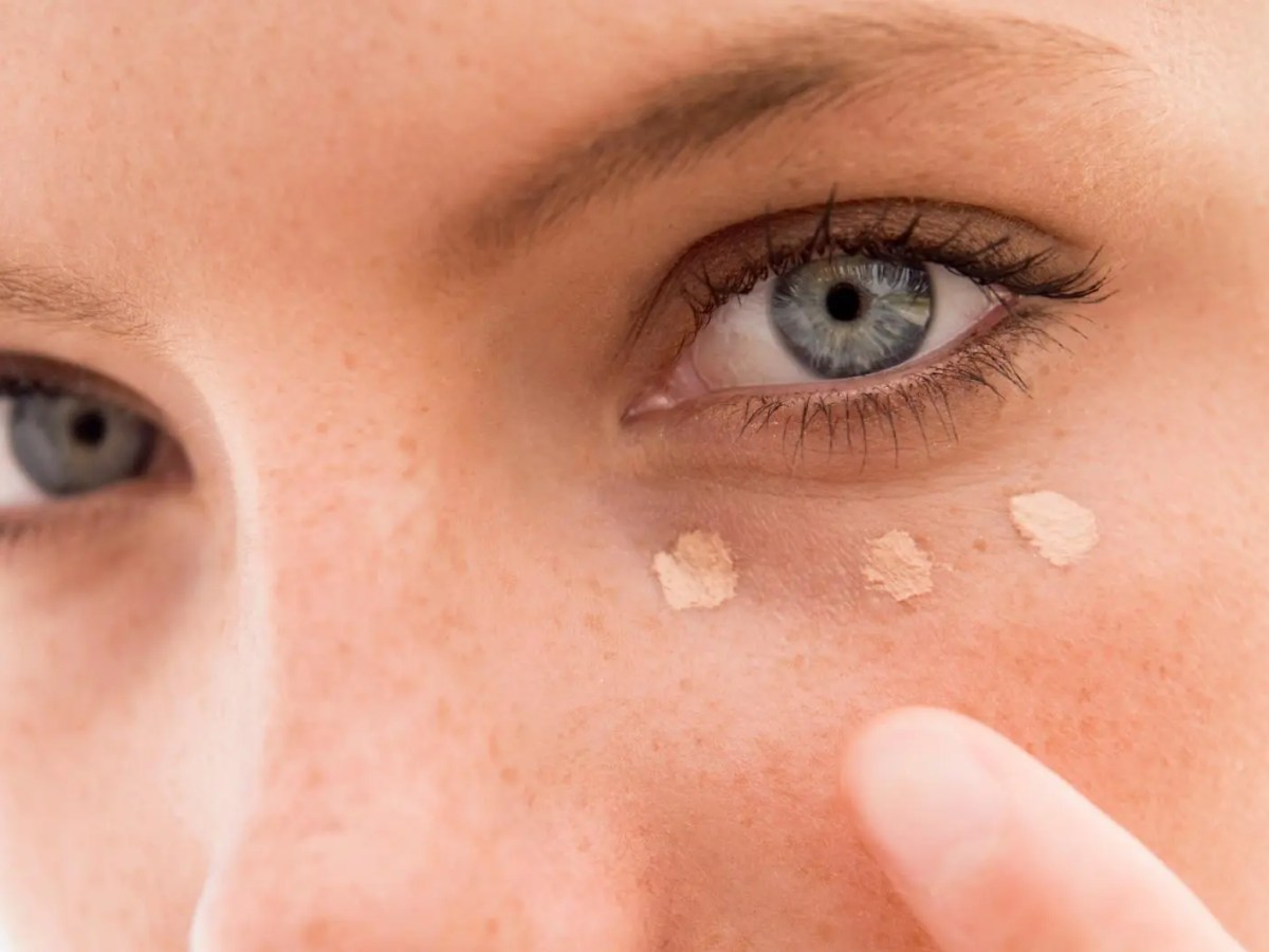 Frau mit hellen blauen Augen tupft sich Concealer unter das Auge.