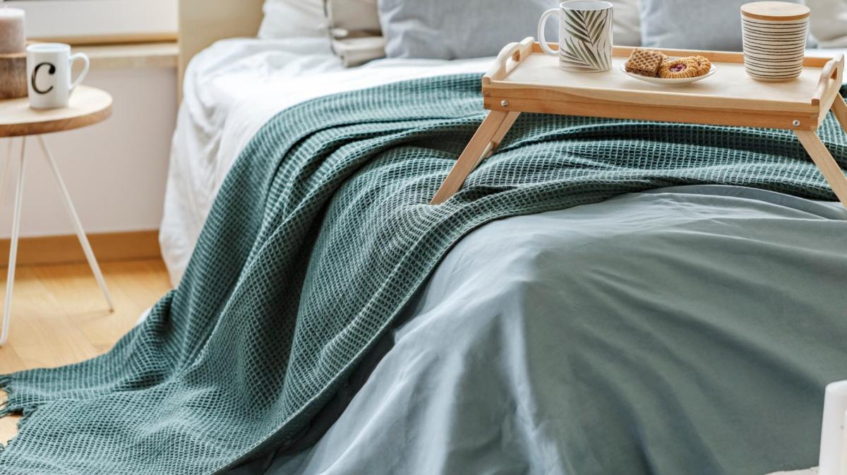 Auf einem Bett liegt eine grün-blaue Tagesdecke und darauf steht ein Tablett mit Tassen und Tellern drauf.