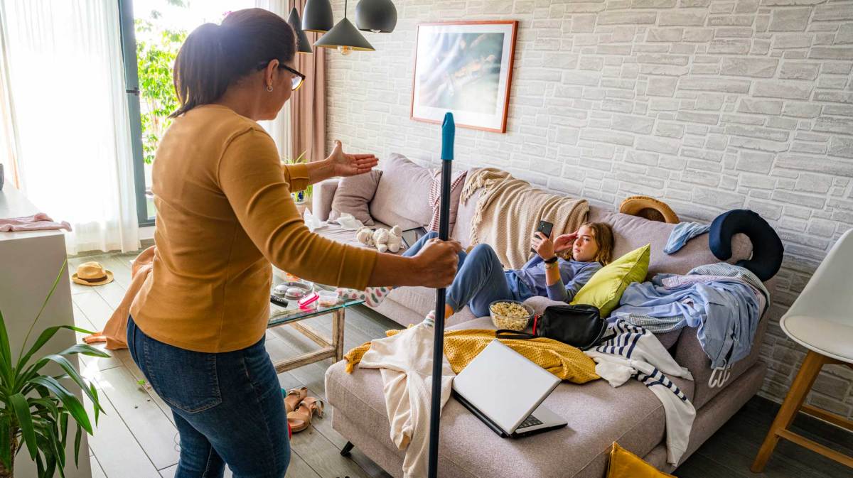 Mutter steht schimpfend vor ihrer Tochter, die im Chaos auf der Couch sitzt und am Handy ist.