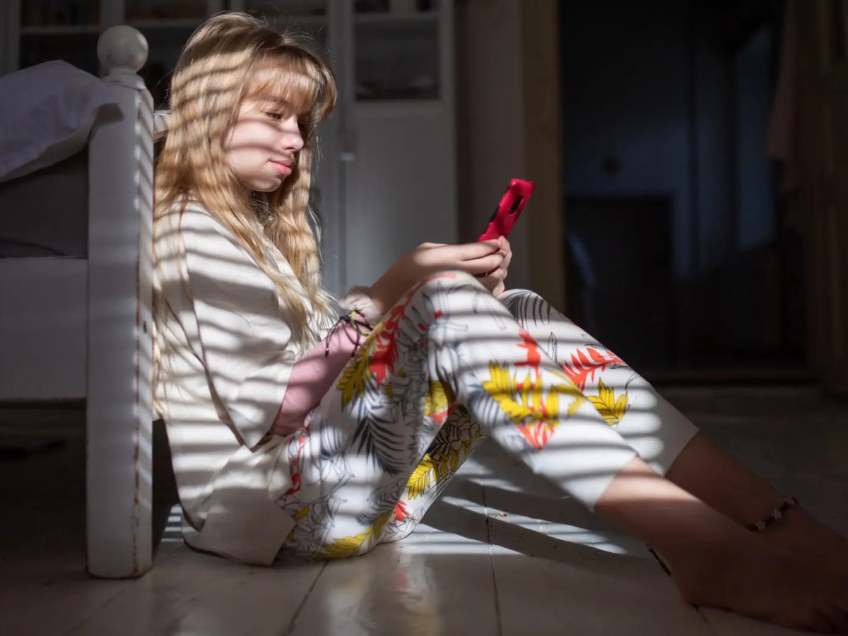 Teenagerin sitzt auf dem Fu&szlig;boden vor ihrem Bett, schaut auf ihr Handy und der Schatten einer Jalousie f&auml;llt auf sie.