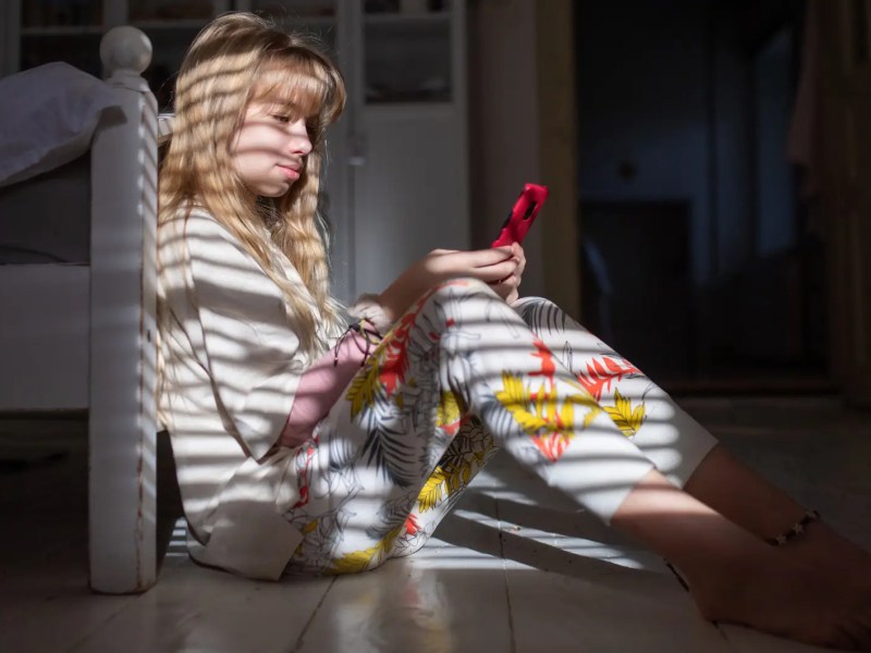 Teenagerin sitzt auf dem Fußboden vor ihrem Bett, schaut auf ihr Handy und der Schatten einer Jalousie fällt auf sie.