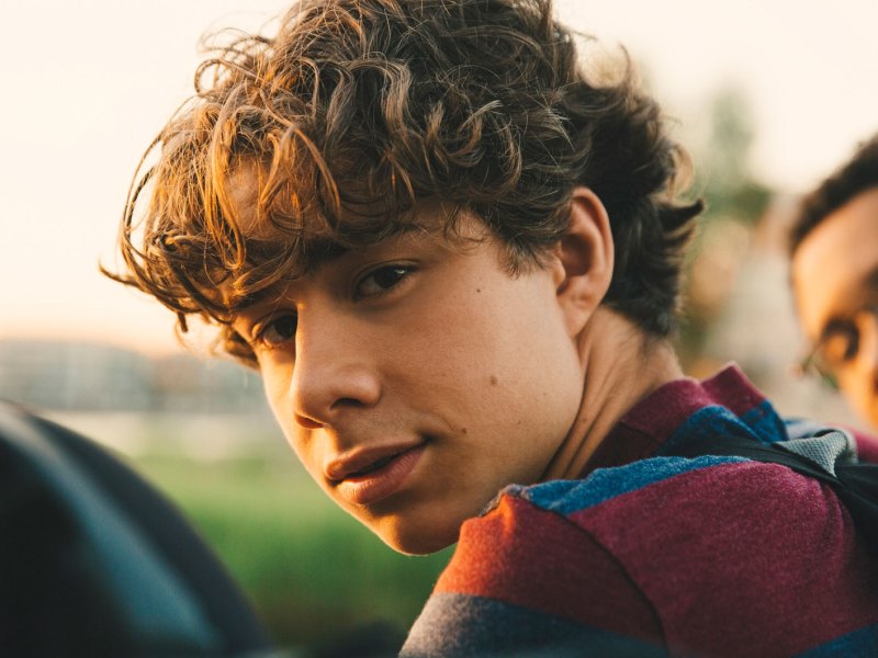 Portr&auml;t eines Teenagers, der drau&szlig;en mit Freunden sitzt und ernst &uuml;ber die Schulter in die Kamera schaut.